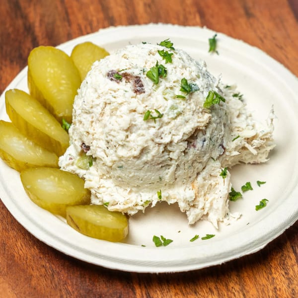 A scoop of Hovan Gourmet Mediterranean chicken salad with parsley and pickle slices on a white plate.